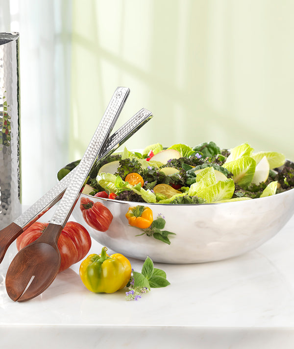 The Mary Jurek Luna Serving Bowl 12" is filled with leafy green salad and vegetables, placed on a marble surface beside wooden salad tongs, fresh tomatoes, and herbs.