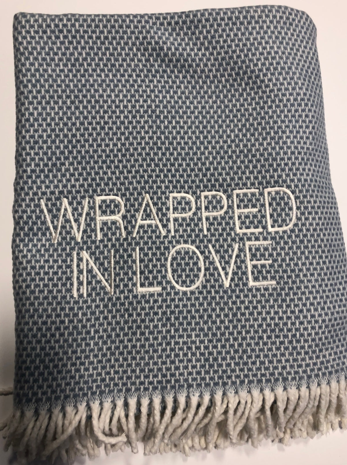 The A Soft Idea Wrapped in Love Throw is a blue and white woven blanket with fringe and “WRAPPED IN LOVE” embroidered in white. Super soft, machine washable, and folded neatly—perfect for gifting.