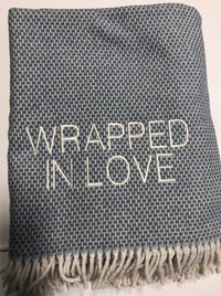 The A Soft Idea Wrapped in Love Throw is a blue and white woven blanket with fringe and “WRAPPED IN LOVE” embroidered in white. Super soft, machine washable, and folded neatly—perfect for gifting.
