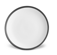 The Soie Tressée Black Dinnerware by L'Objet features a round, white ceramic plate with a dark braided rim, echoing luxury porcelain styles, displayed on a plain white background.