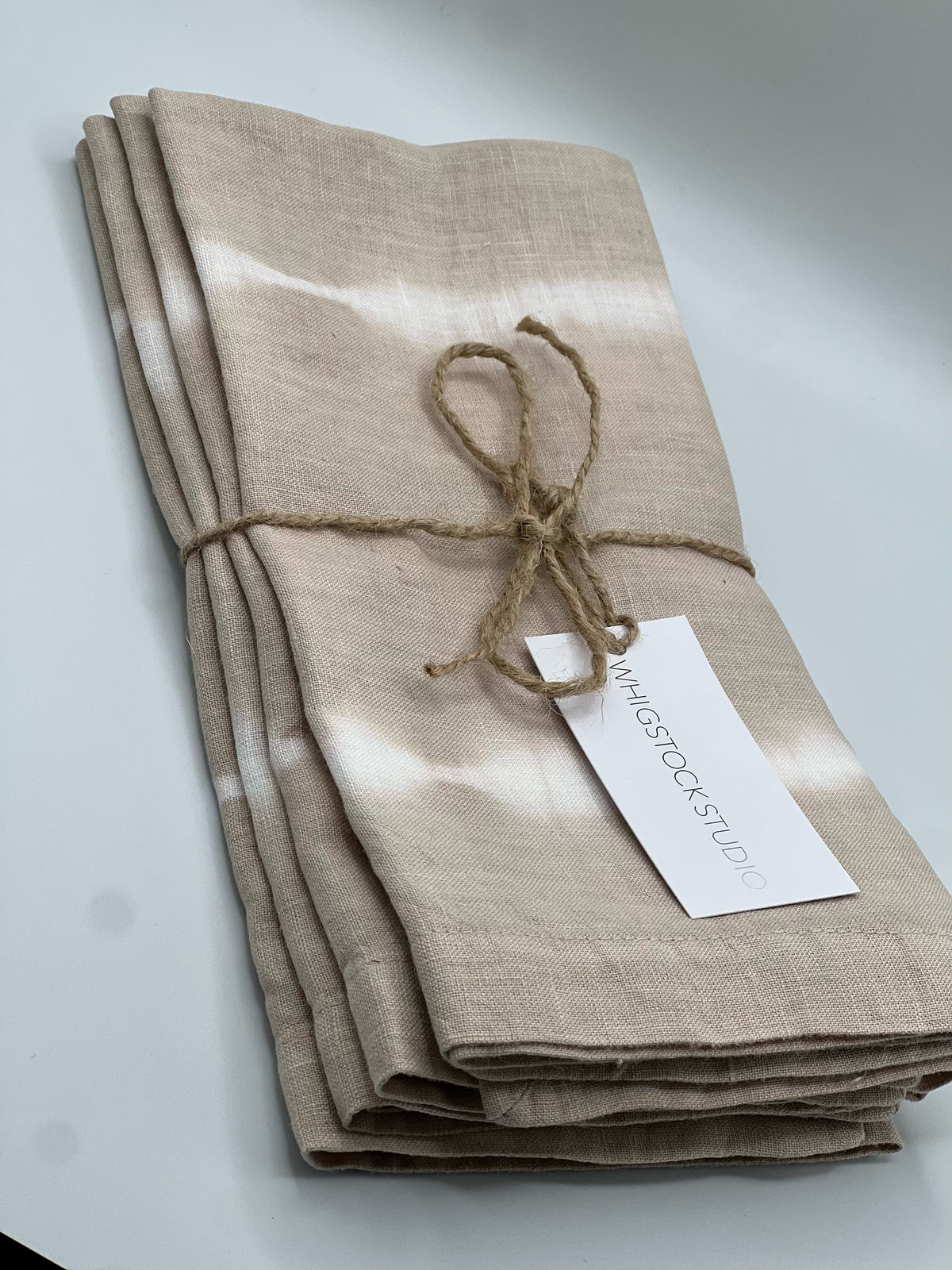 The Whigstock Studio Hand Dyed Linen Napkin Set of 4 - Bone features beige napkins with white tie-dye stripes, neatly tied with twine and a tag—an ideal sustainable essential for any home.
