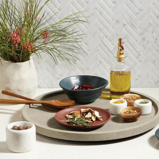 The Calvin Faux Shagreen Lazy Susan Sand by Blue Pheasant serves as a chic centerpiece, displaying dried mushrooms, herbs, oils, and spices—ideal for entertaining. A vase with greenery sits behind on herringbone tile.