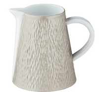 The Raynaud Mineral Irise Dinnerware in Pearl Grey features a textured ceramic jug with a light beige finish, narrow spout, and white handle, reminiscent of Limoges porcelain, set against a plain background.
