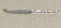 The Sabre Dune Bistrot Cheese Knife Ivory features a curved blade, forked tip, and an ivory handle with a silver swirl design, evoking French flatware elegance. Shown resting on a textured beige surface.