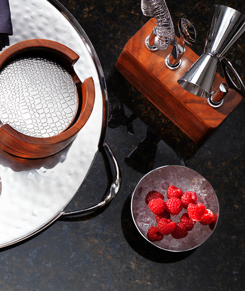 A metal tray holds a wooden and metal coaster holder near a wooden bar tool set with jigger; beside them, a Mary Jurek Luna Serving Bowl 8" is filled with ice and fresh raspberries on a dark countertop.
