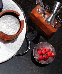 A metal tray holds a wooden and metal coaster holder near a wooden bar tool set with jigger; beside them, a Mary Jurek Luna Serving Bowl 8" is filled with ice and fresh raspberries on a dark countertop.