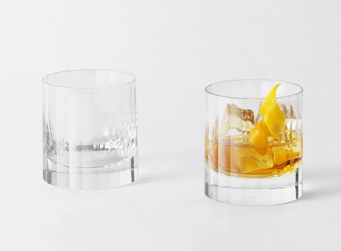 Two elegant Fluted Double Old Fashioned tumblers by Richard Brendon sit on a white surface; the left is empty, while the right contains an amber drink, ice cubes, and a twist of lemon peel.