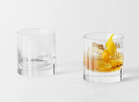 Two elegant Fluted Double Old Fashioned tumblers by Richard Brendon sit on a white surface; the left is empty, while the right contains an amber drink, ice cubes, and a twist of lemon peel.