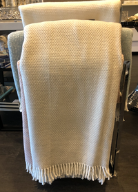 The Grain of Rice Throw by A Soft Idea, in a cream shade and featuring soft fringe, is draped neatly over the back of a light blue chair in a modern room.
