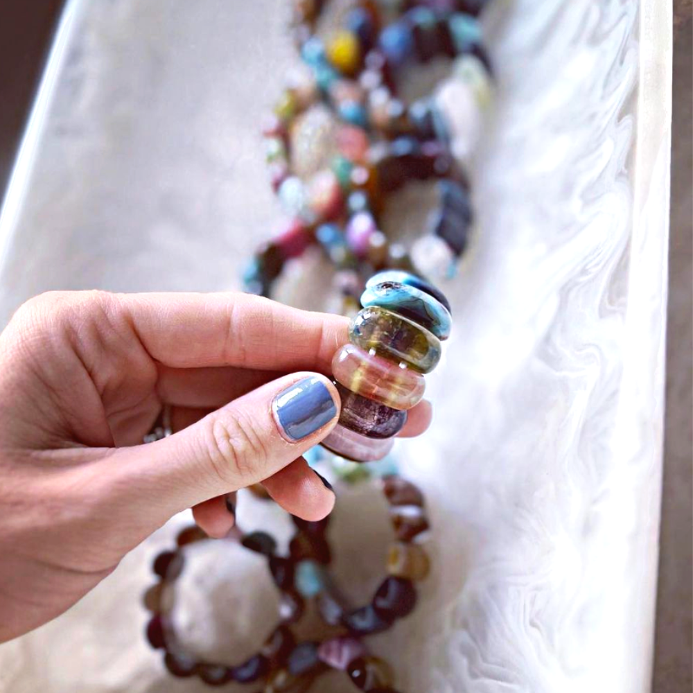 A hand with blue painted nails holds vibrant, rounded glass beads. In the background, beaded jewelry and the Whigstock Studio Gemstone Napkin Ring Multi Set of 4 sit elegantly on a white marbled tray.