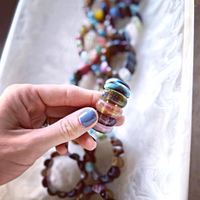 A hand with blue painted nails holds vibrant, rounded glass beads. In the background, beaded jewelry and the Whigstock Studio Gemstone Napkin Ring Multi Set of 4 sit elegantly on a white marbled tray.