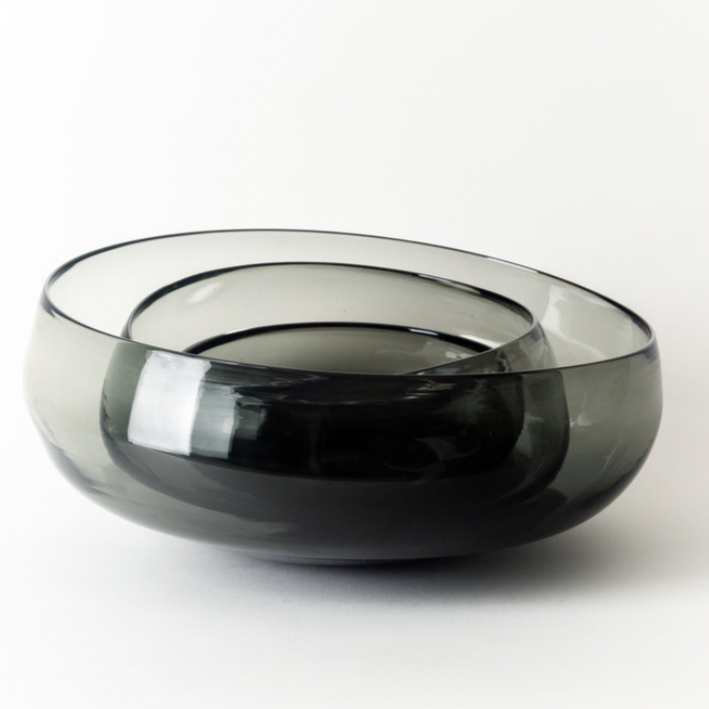 The Gary Bodker Nesting Glass Bowl Charcoal features a modern, smoky gray hand-blown design with a smooth, rounded shape and organic edge, highlighting handcrafted elegance against a plain white background.