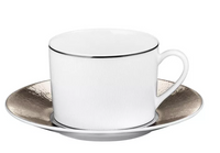 The Bernardaud Dune Dinnerware teacup in white Limoges porcelain features a thin black rim and rests on a matching saucer with a metallic-textured edge, both displayed against a plain white background.