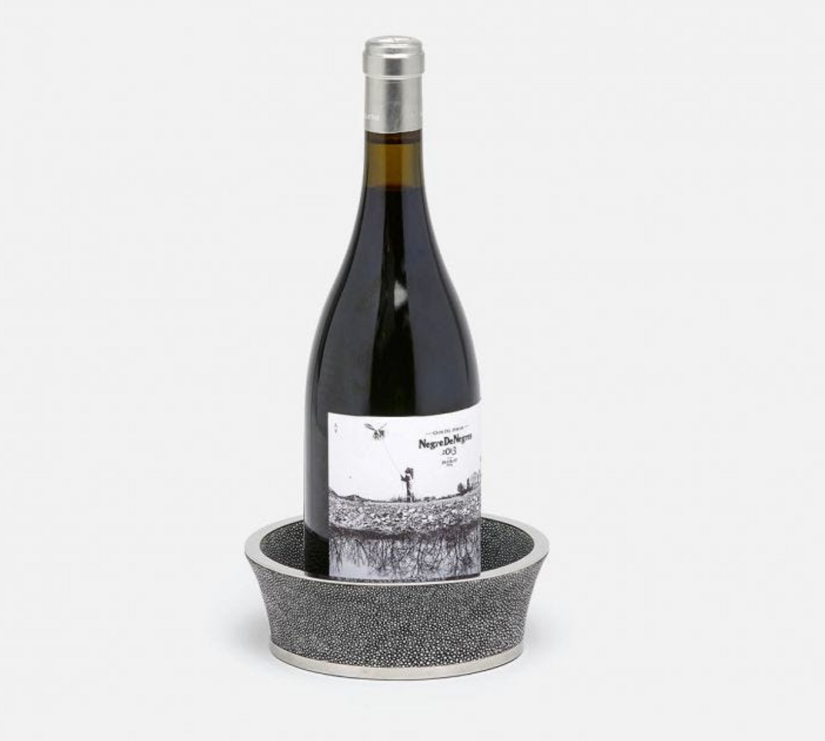 A wine bottle with a white label stands upright in the Angelica Grey Shagreen Wine Coaster by Blue Pheasant, featuring black and silver textured detailing, set against a plain white background.