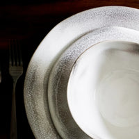 Three overlapping Vietri Aurora Dinnerware Ash pieces—a dinner plate, salad plate, and bowl—showcase a textured gradient gray-and-white stoneware finish. A fork and knife rest on the dark table beside the plates.