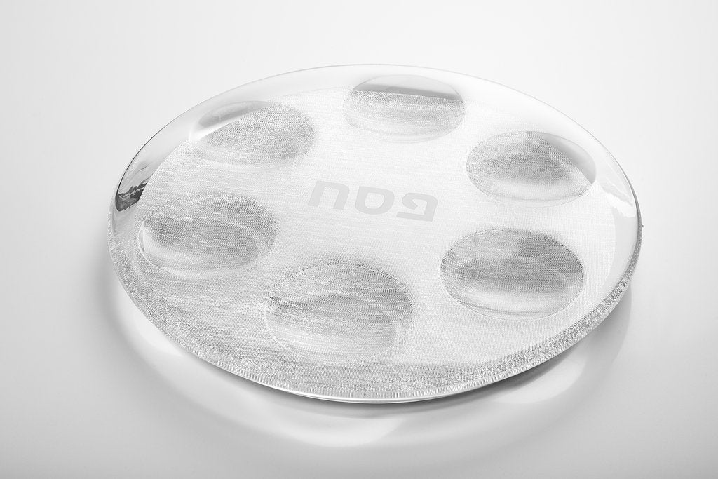 The Apeloig Acrylic Seder Plate is a transparent, round plate with six shallow circular indentations and a modern design, displayed on a white surface. The word "gau" appears faintly in reverse on the plate.