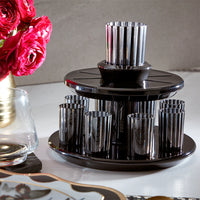 The Apeloig Kiddush Wine Fountain Set, featuring sleek black and silver tones and multiple metallic glasses on a round tray, is displayed on a table beside a glass, red flowers in a gold vase, and a decorative tray.