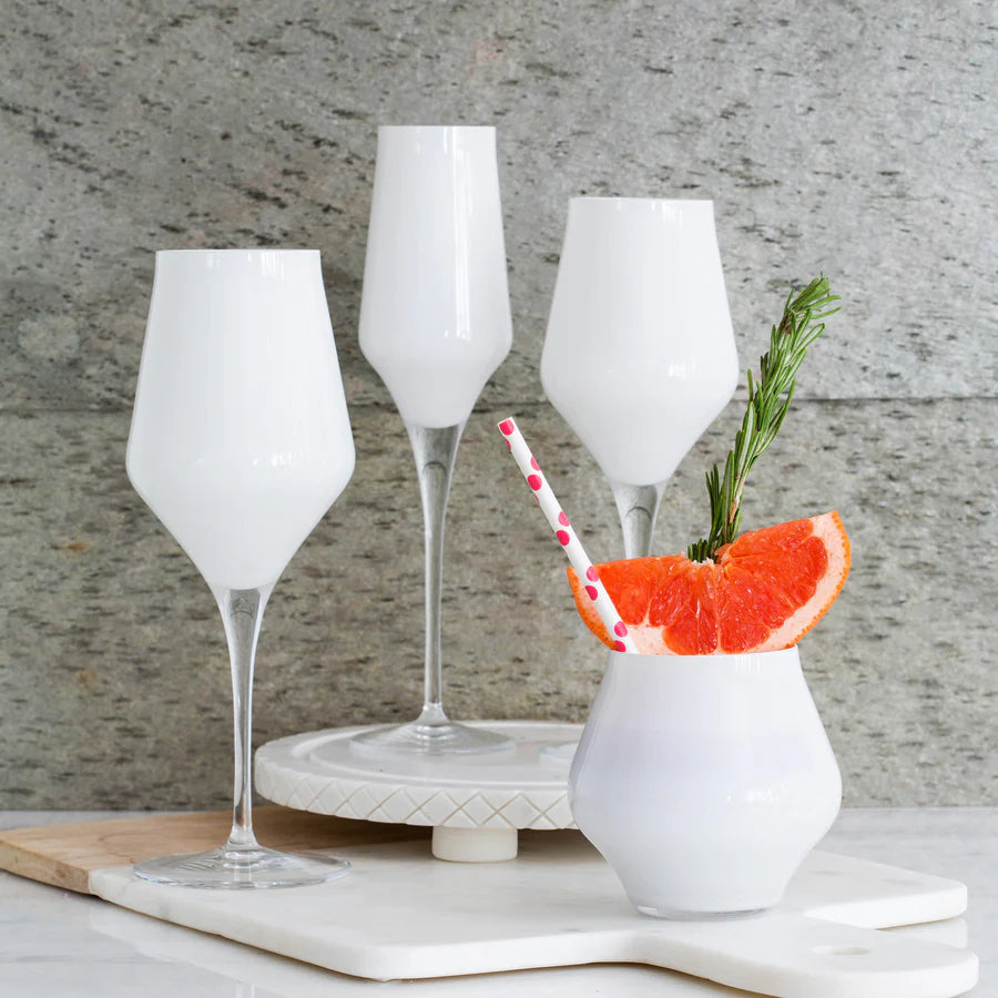 Four white drinks, garnished with grapefruit, rosemary, and a pink straw, are served in elegant Vietri Contessa Stemless Wine Glass White on a marble surface before a stone wall.