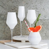 Four white drinks, garnished with grapefruit, rosemary, and a pink straw, are served in elegant Vietri Contessa Stemless Wine Glass White on a marble surface before a stone wall.