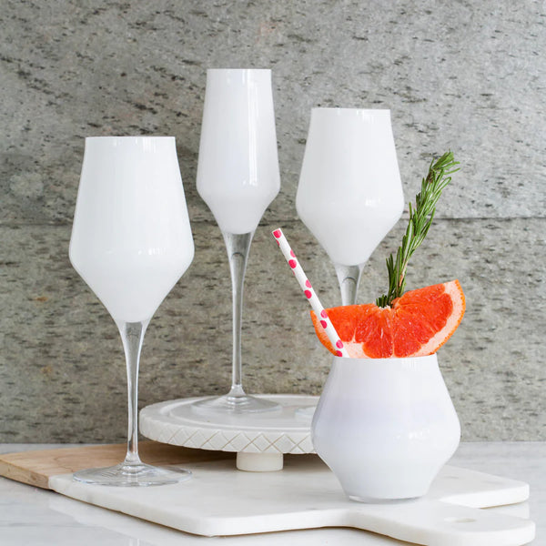 Four white drinks, garnished with grapefruit, rosemary, and a pink straw, are served in elegant Vietri Contessa Stemless Wine Glass White on a marble surface before a stone wall.
