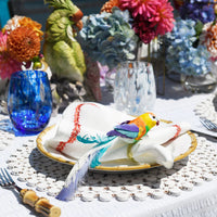 A colorful bird figurine rests on a napkin among vibrant flowers, decorative tableware, and Kim Seybert’s Gala Stemless Tumbler Set 4 on a sunlit outdoor table.