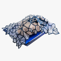 The Geometric Challah Cover by Apeloig features a rock-like sculpted form partially draped in a blue, geometric web-like fabric, resting on a blue rectangular base against a white background.