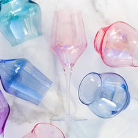 A pink wine glass stands on marble, surrounded by Vietri Contessa Water Glass tumblers—including the Blue—each a vibrant piece of Italian glassware made in Italy.