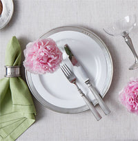 A MATCH Convivio white plate with a pewter rim is set with a silver fork and knife, topped with a pink flower. Nearby are a green cloth napkin in a silver ring and a glass, all on a light tablecloth.