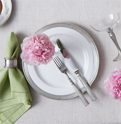 A MATCH Convivio white plate with a pewter rim is set with a silver fork and knife, topped with a pink flower. Nearby are a green cloth napkin in a silver ring and a glass, all on a light tablecloth.