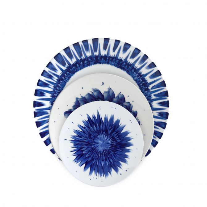 Four white plates from the Bernardaud In Bloom Dinnerware collection are stacked, each showcasing unique blue radial patterns reminiscent of abstract florals or starbursts.