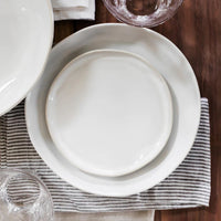 The Forma Cloud Dinnerware by Current Home NY features white stoneware plates, large and small, stacked on a striped gray and white napkin atop a wooden table with clear glassware. Chip resistant for everyday use.