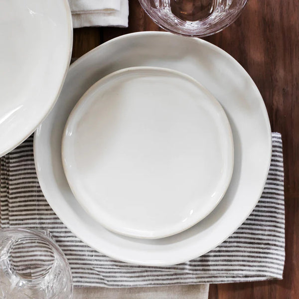 The Forma Cloud Dinnerware by Current Home NY features white stoneware plates, large and small, stacked on a striped gray and white napkin atop a wooden table with clear glassware. Chip resistant for everyday use.