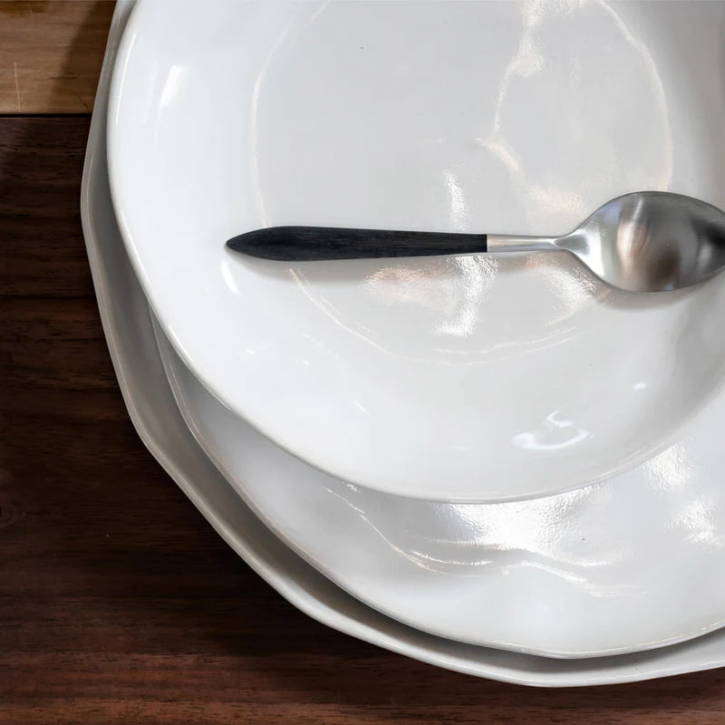 A silver spoon with a dark handle rests on a stack of three Current Home NY Forma Cloud Dinnerware plates, arranged on a dark wooden surface.