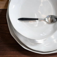 A silver spoon with a dark handle rests on a stack of three Current Home NY Forma Cloud Dinnerware plates, arranged on a dark wooden surface.