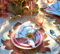 Elegant tablescape featuring pink plates, gold flatware, a white napkin adorned with the stunning Kim Seybert Stellar Napkin Ring Gold Set of 4, pink glasses, and autumn-themed placemats. Decorative accents include a gold ant figurine and a floral centerpiece.