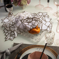 A braided challah loaf partially covered by the Apeloig Geometric Challah Cover, featuring Hebrew text, surrounded by elegant glassware, plates, and napkins set for a meal.