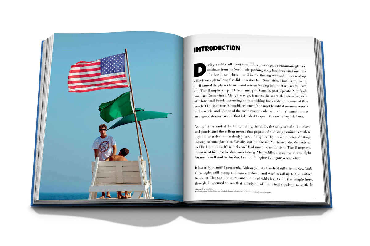 An open copy of Hamptons Private by Assouline shows a photo of two people on a lifeguard chair with American and green flags under a blue sky. The opposite page features text titled INTRODUCTION, evoking summer in the Hamptons.