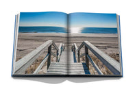 An open book, "Hamptons Private" by Assouline, displays a photo of wooden stairs to a sandy beach and calm ocean, capturing the serene beauty and inviting summer escape vibes of The Hamptons.