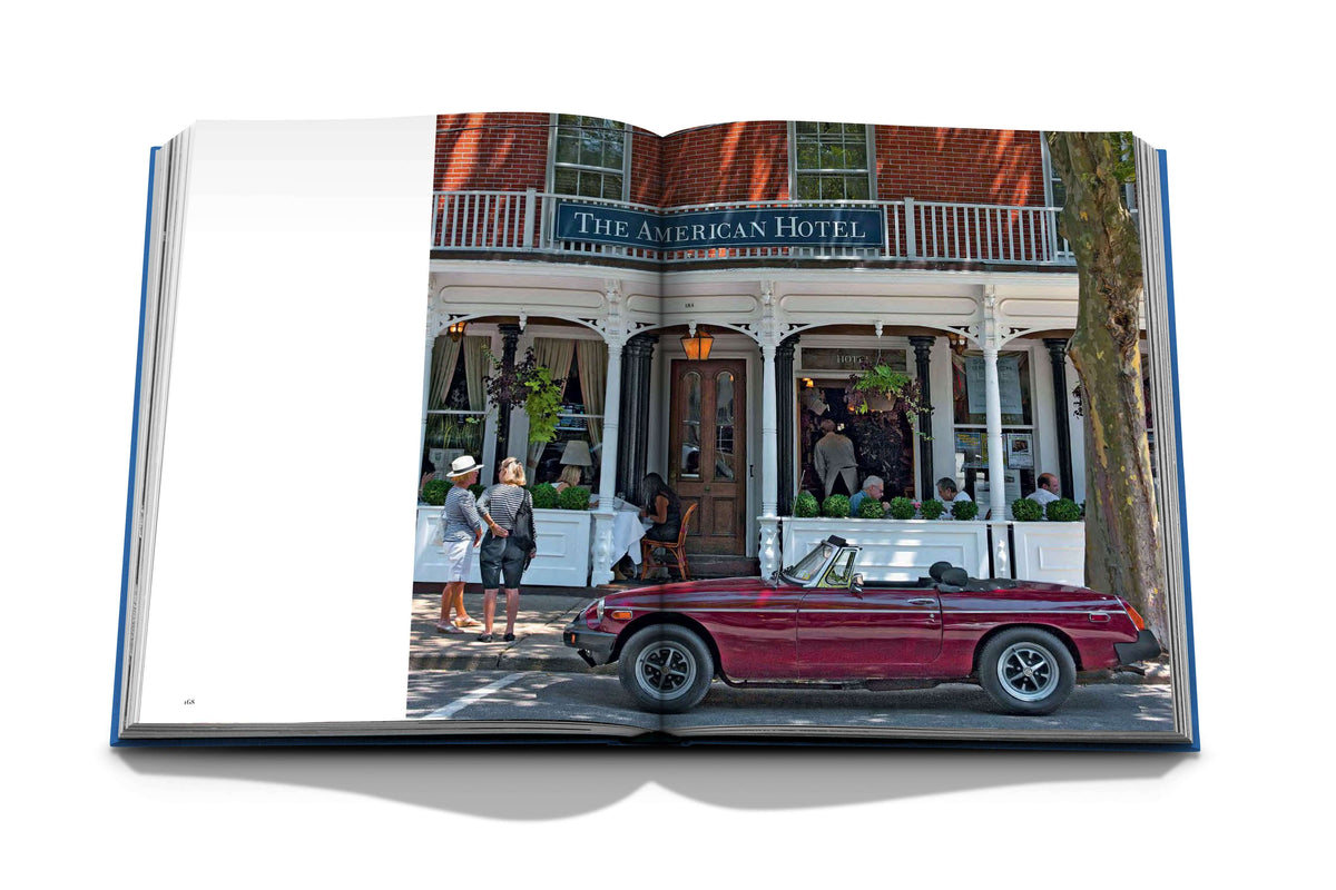 An open book, Hamptons Private by Assouline, features a photo of The American Hotel in The Hamptons—a red vintage convertible out front and people on the porch—evoking summer charm along these iconic luxury real estate-lined streets.