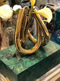 The Trui Vase by Rosenthal, featuring a shiny gold looped design and holding two white calla lilies, sits atop a green marble-patterned box. Decorative objects and flowers decorate the blurred background, enhancing its elegant appeal.