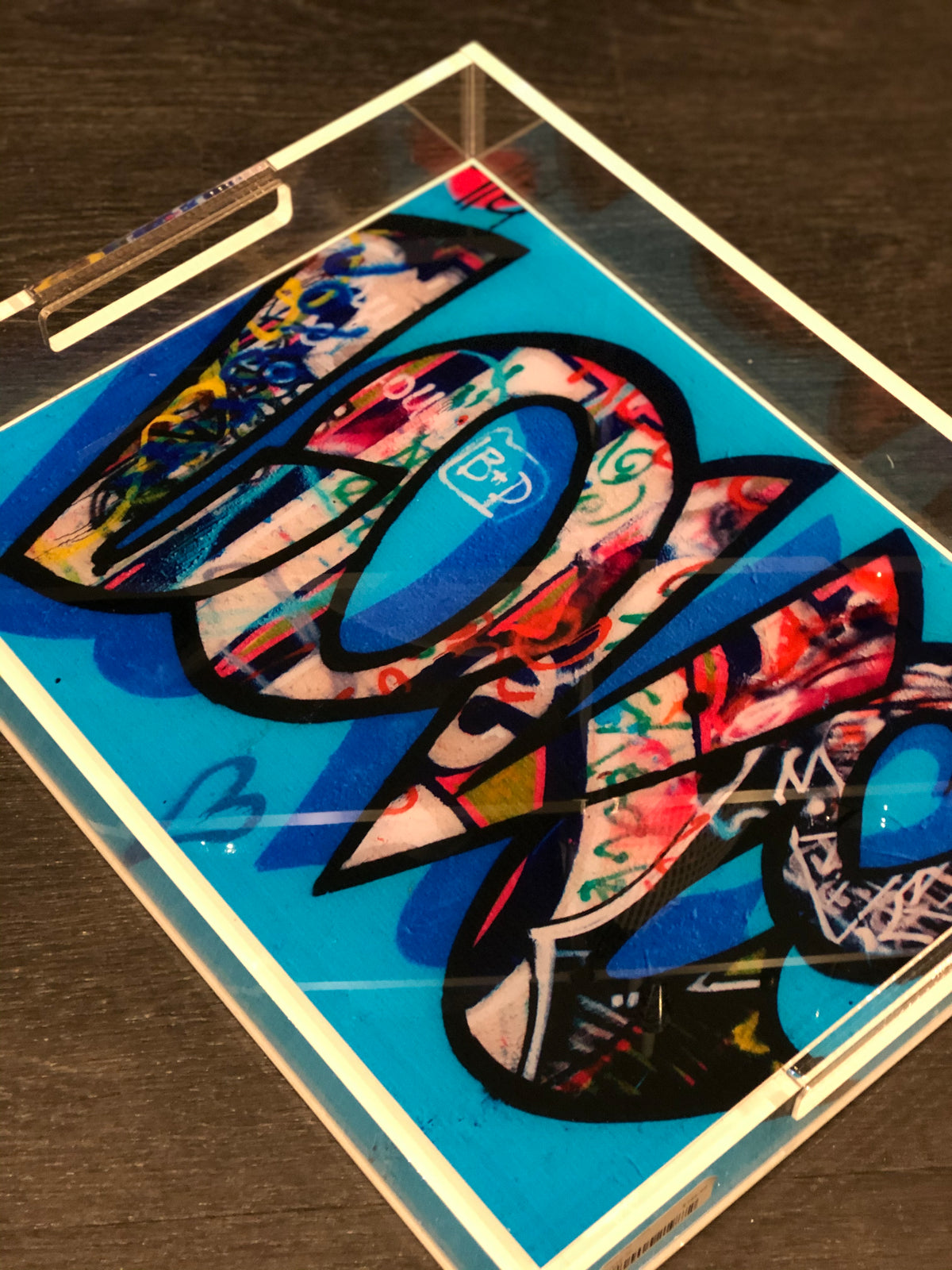 The Eye Candy Graffiti Love Tray | Limited Edition features Chelsea Beck's vibrant graffiti-style "LOVE" art on clear acrylic with a bold blue background, shown on a dark wooden surface.