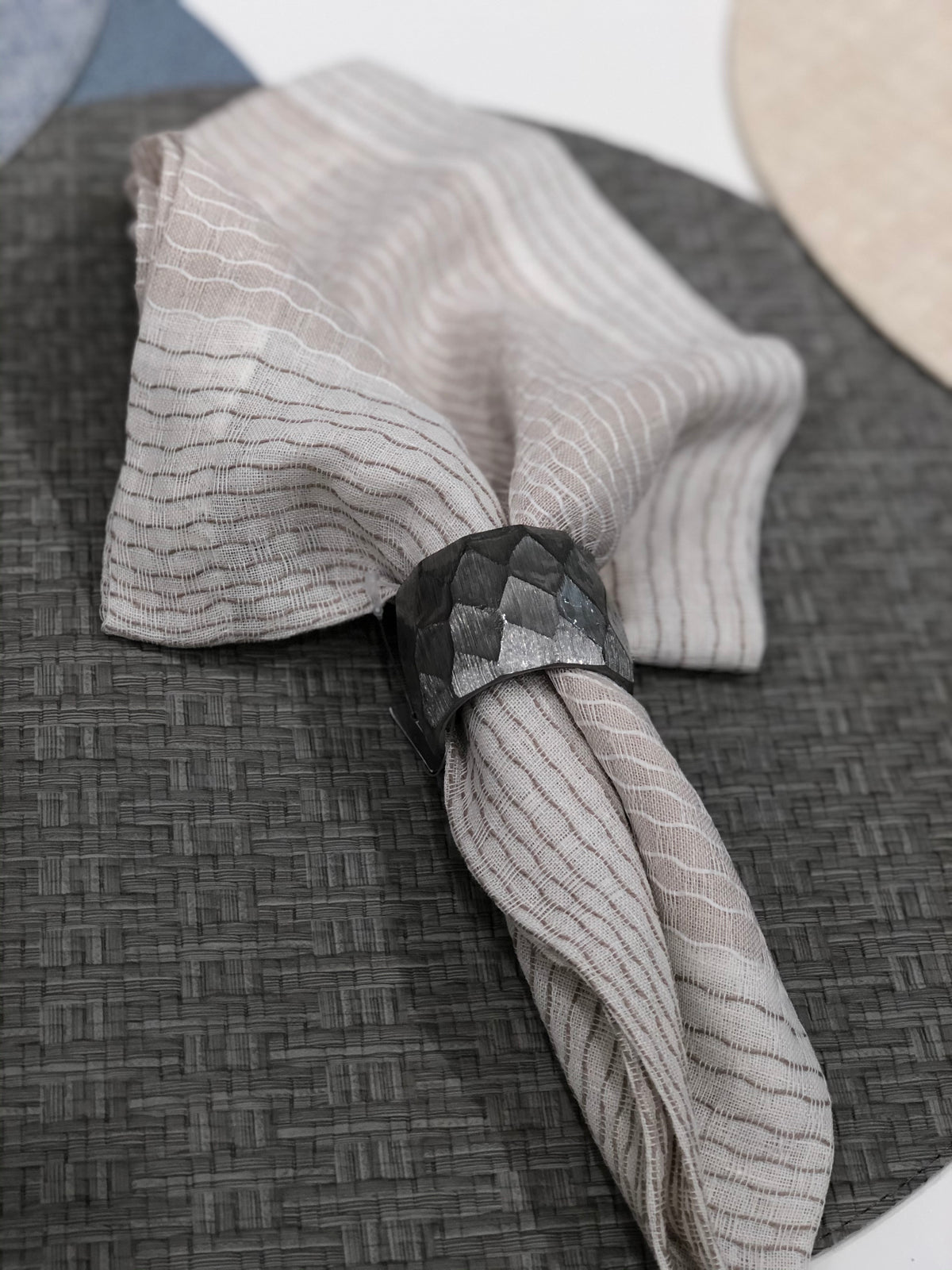 A white napkin with thin brown stripes is held by a textured, dark metallic napkin ring and placed on Bodrum's Wicker Round Placemat Grey (Set of 4), offering stylish, easy-care elegance to any table setting.