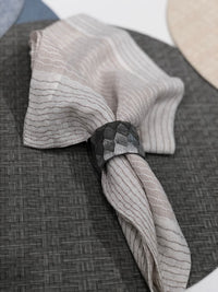 A white napkin with thin brown stripes is held by a textured, dark metallic napkin ring and placed on Bodrum's Wicker Round Placemat Grey (Set of 4), offering stylish, easy-care elegance to any table setting.