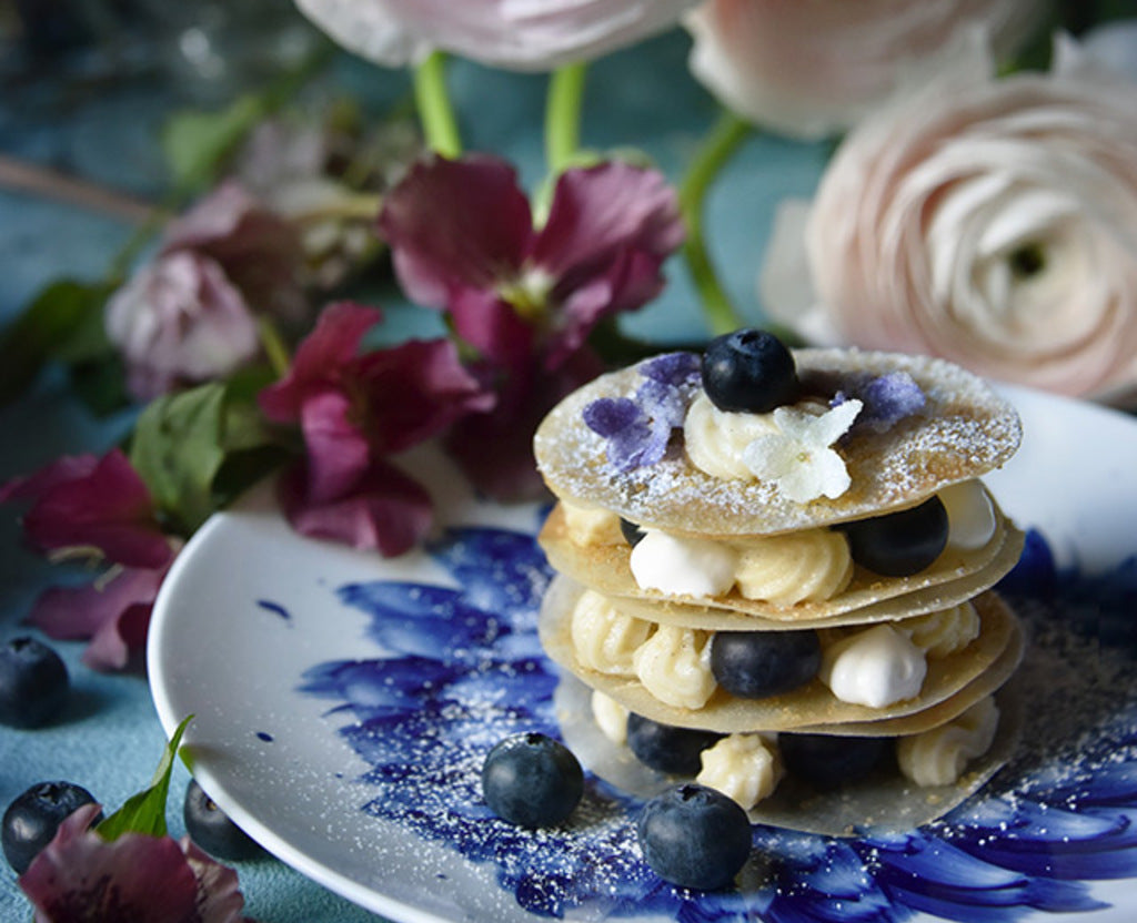 A dessert with three pastry layers, cream, blueberries, powdered sugar, and edible flowers sits on Bernardaud’s In Bloom Dinnerware—a plate adorned with blue brushstroke designs and pink flowers from the porcelain collection.