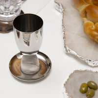 The Anna New York Coluna Marble Kiddush Cup Silver, with a modern design, rests on its saucer next to braided challah, green olives in a dish, and a glass of water on a white table.