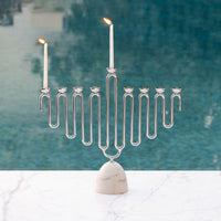 The Coluna Menorah Carrara Marble & Silver by Anna New York features a modern silver menorah with two lit candles standing elegantly on a white Italian Carrara marble base, set against a softly blurred blue background for a timeless display.