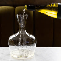 A hand pours white wine from a green bottle into a lead-free crystal Jancis Robinson Young Wine Decanter by Richard Brendon on white marble, with a blurred dark background.