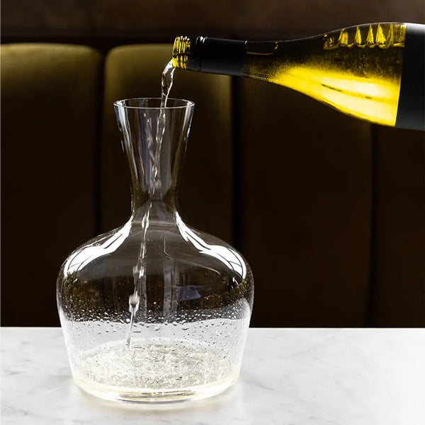 A hand pours white wine from a green bottle into a lead-free crystal Jancis Robinson Young Wine Decanter by Richard Brendon on white marble, with a blurred dark background.