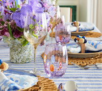 A close-up of a table with a striped blue and white cloth, gold-rimmed wine glass, Kim Seybert's Gala Stemless Tumbler Set of 4, woven placemats, blue napkins with rings, and purple flowers in the background.