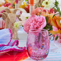 The Kim Seybert Gala Stemless Tumbler Set 4, featuring colorful glass with a pink swirl pattern, is displayed on a striped tablecloth with bright flowers, woven bunny napkin rings, and vibrant pink-trimmed napkins.